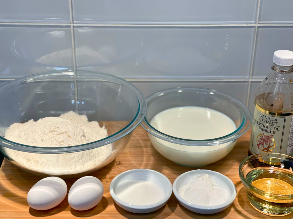 ingredients to make waffles batter at home including flour, milk, eggs, oil, vanilla, sugar, and corn starch