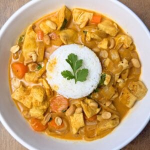 A bowl of creamy coconut chicken curry with orange carrots, served next to white basmati rice and topped with crushed peanuts and fresh green coriander.